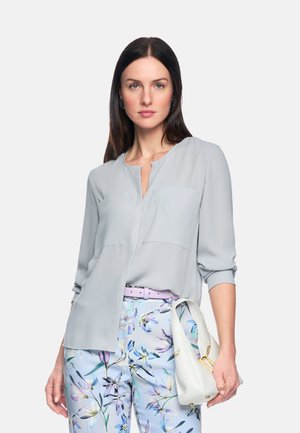 Frau mit langem dunklem Haar, die eine hellgraue Bluse und eine Hose mit Blumenmuster trägt, hält eine weiße Handtasche vor einfarbigem Hintergrund.