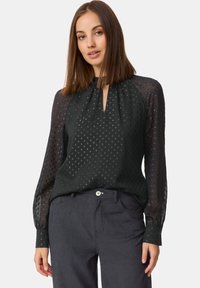 Schwarze Bluse mit langen Ärmeln, gewebtem Punktemuster, Schlüsselloch-Ausschnitt und transparenten Ärmeln. Kombiniert mit navyblauen Cordhosen.