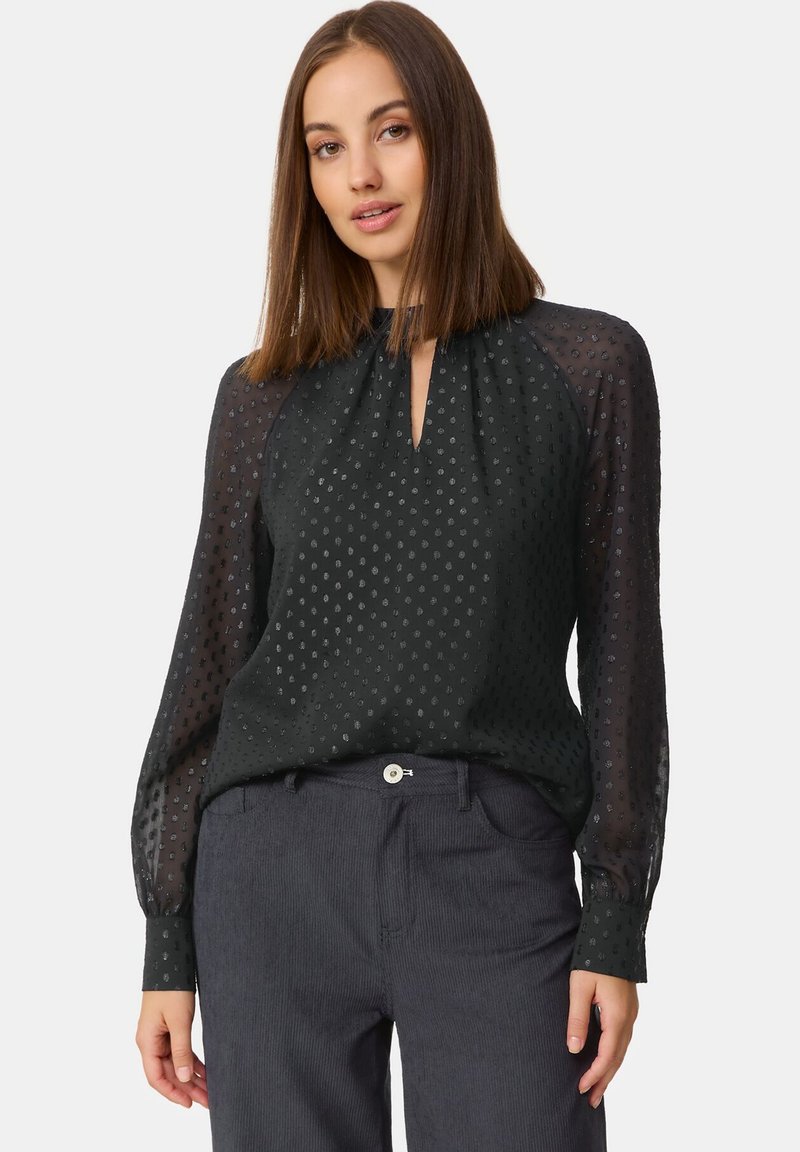 Schwarze Bluse mit langen Ärmeln, gewebtem Punktemuster, Schlüsselloch-Ausschnitt und transparenten Ärmeln. Kombiniert mit navyblauen Cordhosen.