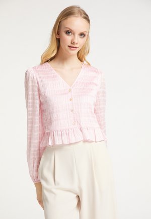 Jonge vrouw met lang blond haar die een lichtroze V-hals blouse met knopen en een gerimpelde zoom draagt, gecombineerd met hooggetailleerde crèmekleurige broek.