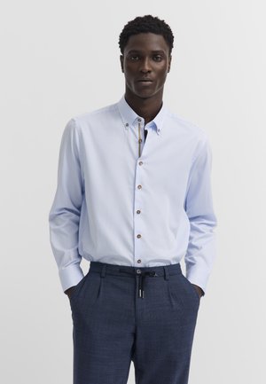 Mann trägt ein hellblau gestreiftes Hemd, in dunkelblaue, maßgeschneiderte Hose gesteckt, mit den Händen in den Taschen, vor schlichtem Hintergrund stehend.