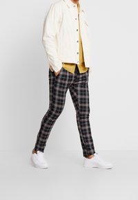 Svarta och vita rutiga byxor kombinerade med en gul skjorta och en krämfärgad denimjacka, fulländat med vita sneakers.