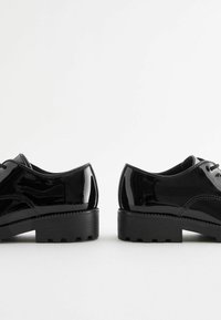 Chaussures en cuir verni noir avec un bout arrondi, un laçage et une semelle épaisse en caoutchouc noir texturé. Fini lisse et brillant.