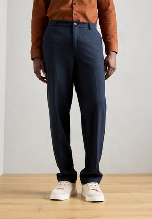 Pantaloni blu navy con vestibilità tailored, caratterizzati da una vita piatta, passanti per la cintura e una chiusura con un solo bottone. Indossati con sneaker beige.