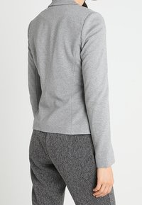 Blazer ajustado gris con una textura suave, cuello estructurado y mangas largas. Combinado con pantalones oscuros y con patrón. Se muestra vista trasera.