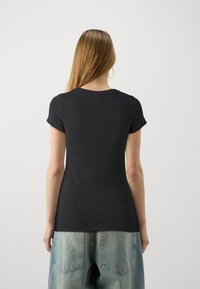 Žena s dlouhými vlasy oblečená v černém přiléhavém tričku s krátkým rukávem a světle modrých džínech, stojící zády k jednoduchému pozadí.