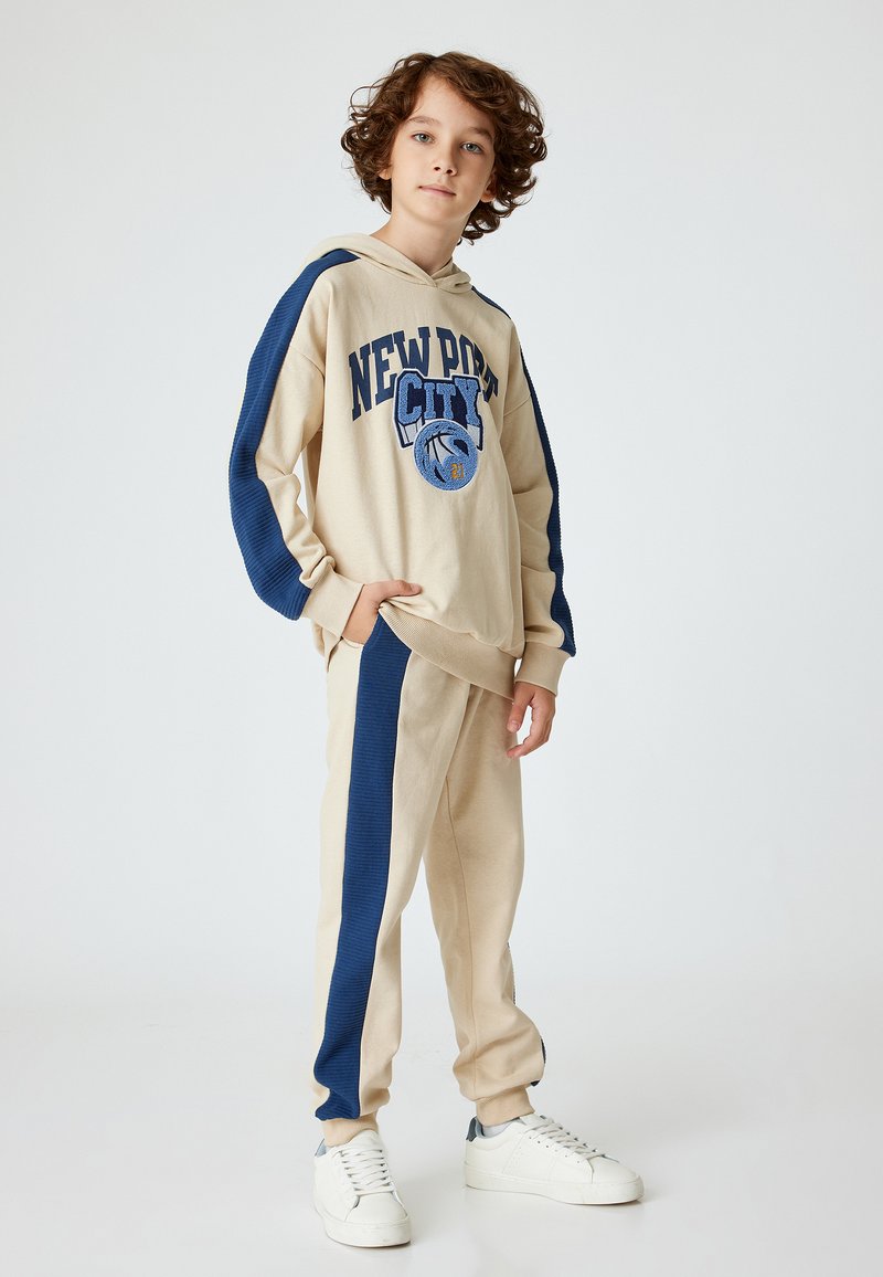 Beige hoodie met marineblauwe accenten, voorzien van een "NEWPORT CITY" graphic. Gecombineerd met beige joggers, ook met marineblauwe zijpanelen en een cuffed enkel.