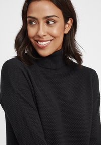 Schwarzer Strickpullover mit hohem Kragen, der ein strukturiertes Muster und eine lockere Passform aufweist. Trägt eine Person mit schulterlangem dunklem Haar.