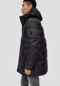 Chaqueta de abrigo negra con capucha, con un diseño acolchado, bolsillos laterales y un logo en la manga. Hecha de un material brillante y acolchado.