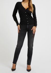 Black cardigan with a V-neck and button front, paired with dark jeans featuring ornate lace detailing, and pointed black heels.