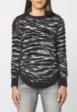 Femme portant un pull tricoté rayé en noir et blanc, doux, à manches longues, et un jean gris clair, debout devant un fond uni.