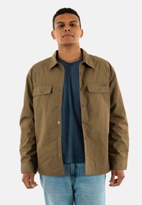 Camicia button-up marrone con due tasche sul petto, realizzata in materiale cotone. Indossata sopra una t-shirt blu a scollo a V, abbinata a jeans chiari.