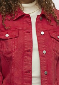 Rød denimjakke med knaplukning foran, to brystlommer og sølvfarvet hardware. Har en klassisk krave og en let struktureret overflade.