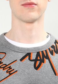 Ingrandimento della parte inferiore del viso e del collo di una persona che indossa un maglione grigio con un design di testo astratto arancione e nero, e una camicia bianca sotto.