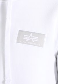 Witte hoodie van een katoenmix, voorzien van een rechthoekig grijs patch met het "Alpha Industries" logo en hoekige ontwerpelementen.