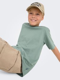 Grøn T-shirt med korte ærmer og afslappet pasform, sammen med beige bukser. Barnet har en beige kasket på med teksten "NEW YORK CITY ATHLETICS".