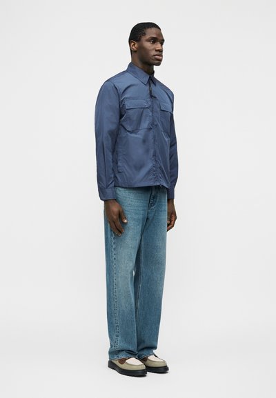 Homme portant une chemise bleu à boutons avec poches poitrine, un jean bleu ample et des chaussures blanches et brunes, debout devant un fond uni.
