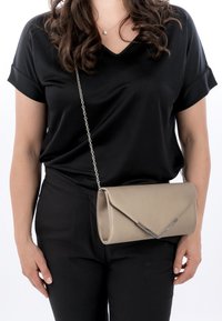 Mujer vestida con un conjunto negro y un bolso de mano beige con un detalle de metal plateado y una correa de cadena, visible desde el pecho medio hasta las caderas.