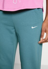 Teal Nike sweatpants med en glat tekstur, som har et broderet hvidt logo. Sammen med en lyserød top, der viser en afslappet pasform.