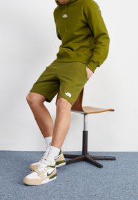 Zelená bavlněná mikina s kapucí a odpovídající šortky s bílým logem. Hnědé a zelené tenisky a bílé ponožky. Sedící na dřevěné židli. Modrý koberec.