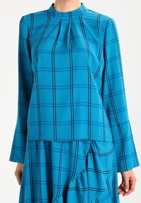 Top turchese con maniche lunghe, caratterizzato da un collo alto e un design plissettato. Motivo a quadri neri su tutta la superficie; tessuto con finitura liscia.