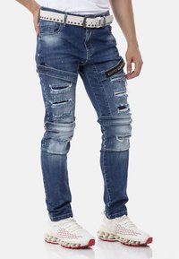 Ausgebleichte blaue Jeans mit beschädigten Stellen und einem schwarzen Logo-Patch, kombiniert mit einem weißen Gürtel und weißen Sneakers mit roten Akzenten.