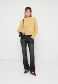 Fluffy gele trui, zwarte tapered jeans en rode ballerina's. Model houdt een kleine zwarte handtas vast. Eenvoudige achtergrond in neutrale kleur.