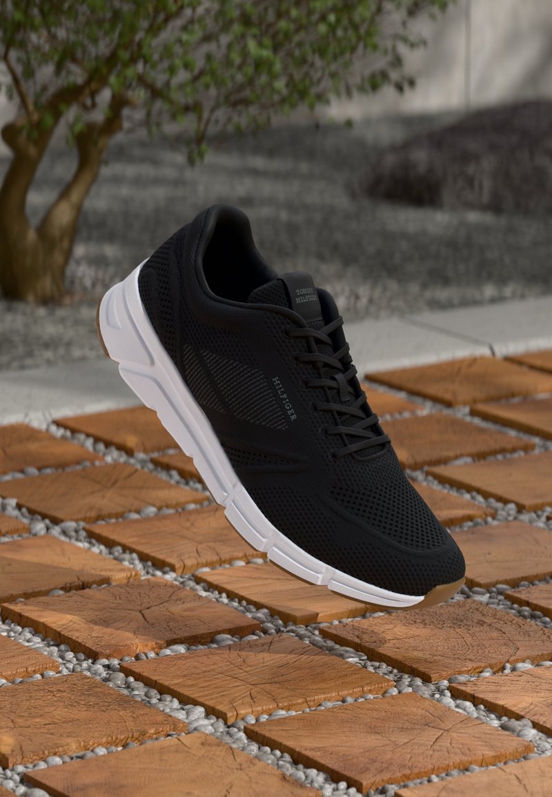 Chaussure de sport noire avec semelle blanche et logo Tom Hilfiger sur le côté, flottant au-dessus de carreaux carrés en bois avec de petits cailloux gris entre eux.