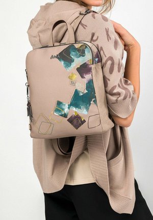 Donna che indossa un maglione beige e tiene una borsa taupe con disegni quadrati astratti blu, viola e oro sulla spalla.