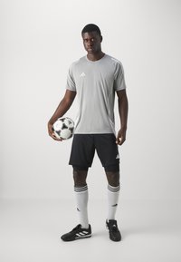 Szürke sportpóló fehér részletekkel, fekete rövidnadrág, fekete focicipő és fehér zokni fekete csíkokkal, egy focilabdát tartva.