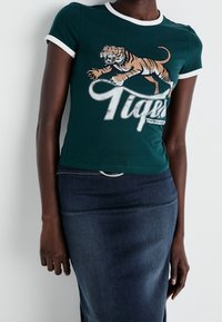 Camiseta de algodón verde con ribetes blancos, que presenta un gráfico de un tigre y el texto "Tigers" en blanco desgastado. Llevada con una falda de mezclilla oscura.