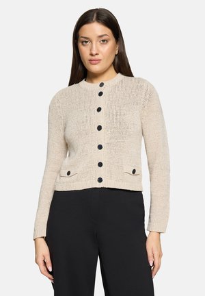 Beżowy dziergany kardigan z okrągłym dekoltem, czarnymi guzikami i dwiema przednimi kieszeniami. Teksturowany wzór; dopasowany fason. Noszony z czarnymi spodniami.