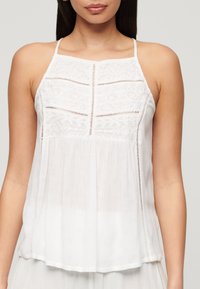 Witte mouwloze top met ingewikkelde kantdetails, een geplooide rok en een lichtgewicht, semi-doorzichtige stof.