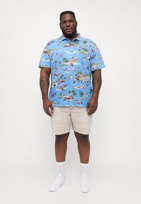 Camisa polo azul de mangas curtas com um padrão tropical que inclui palmeiras, barcos e flores. Usada com calções bege e ténis brancos.