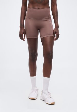 Atletisk person iført brune højtaljede tætsiddende shorts med sidelommer, hvide crew-sokker og hvide løbesko på ensfarvet baggrund.