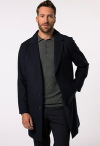 Cappotto di lana blu navy con colletto a punta, chiusura a bottone monopetto e tasche laterali, indossato sopra una maglietta polo grigia e pantaloni.