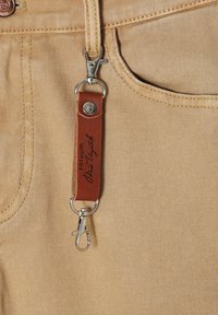 Tela de mezclilla beige con un patrón texturizado, con una correa de cuero marrón con herrajes de plata y texto grabado unido al bolsillo.