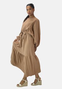 Robe marron à manches longues avec une jupe à volants et une taille ceinturée. Tissu doux, encolure arrondie et manches froncées. Portée avec des sandales compensées dorées.