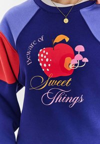 Kolorowy sweatshirt z niebieską bazą, czerwonymi akcentami oraz grafiką przedstawiającą jabłko i grzyby z napisem "Strzeż się słodkich rzeczy."