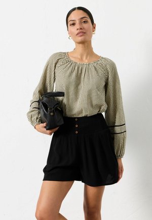 Kvinde iført en beige ternet langærmet bluse og sorte shorts med høj talje, der holder en sort håndtaske mod en hvid baggrund.