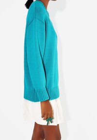 Turkusowy sweter z dzianiny z prążkowanym dołem i mankietami. Ma luźny krój. Noszony na białej spódnicy warstwowej. Zielona obrączka w kształcie kokardy na dłoni.