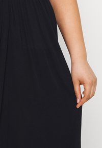 Mano destra che tiene delicatamente il lato di una gonna nera a pieghe su uno sfondo chiaro.