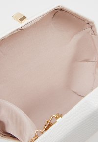 Bílá texturová kabelka na zips se zlatým claspem, s hladkým béžovým interiérem a malým zlatým kroužkem na řetězové připevnění.