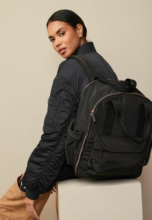 Schwarzer Rucksack mit mehreren Taschen, Reißverschlüssen und verstellbaren Trägern. Kombiniert mit einer schwarzen Bomberjacke mit tonal abgestimmten Nähten und gerippten Bündchen.