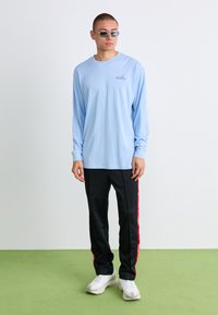 Mand iført en lyseblå langærmet Nike-shirt, sorte bukser med røde sidestriber, hvide sneakers og grønne solbriller, står på et grønt gulv.