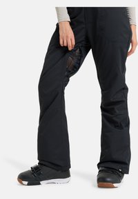 Zwarte skibroek met een strak ontwerp, voorzien van een meshpaneel op de linkerdij en een rits. Gepaard met zwarte snowboardlaarzen.