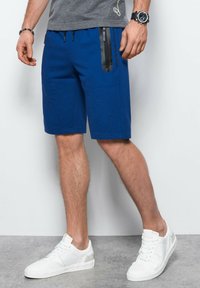 Pantaloni corti blu con una finitura testurizzata, dotati di una tasca con cerniera nera, laccetti laterali e una vestibilità comoda, abbinati a sneaker bianche.