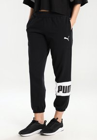 Benen in zwarte Puma joggers met een wit logo op de rechter kuit, gecombineerd met zwarte sportschoenen met witte zolen. Handen in de zakken.