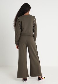 Olivgrön jumpsuit med vida ben, ribbat midjeband och vita knappdetaljer på axlarna. Tillverkad av mjukt material.