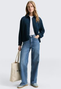 Donkerblauwe corduroyjas, witte shirt, lichtblauwe hoge taille jeans en beige tote bag. Schoenen hebben een lichtblauw bovenwerk en een bruinzolen.